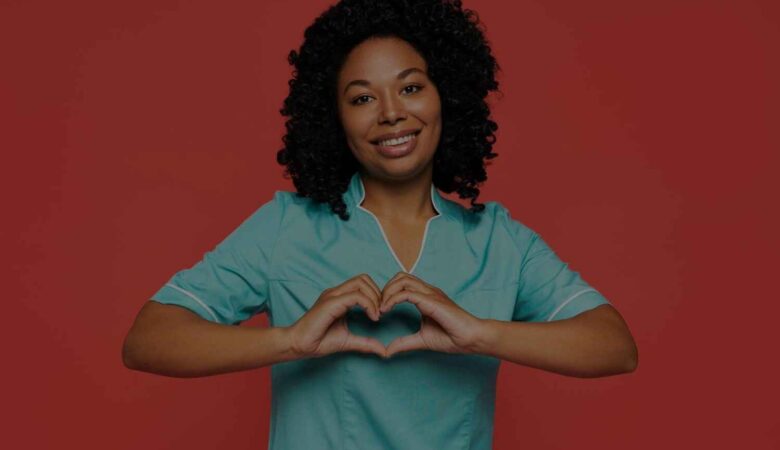 The image shows a smiling healthcare professional wearing a light blue uniform, standing against a solid red background. She is making a heart shape with her hands in front of her chest, symbolizing care and compassion. Her expression is warm and welcoming, and the overall image conveys a sense of empathy, support, and dedication, often associated with those in the healthcare field.