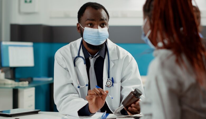 A male doctor, wearing a white coat, stethoscope, and face mask, is seated at a desk in a consultation room. He is holding a bottle of medication and explaining something to a patient, who is also wearing a face mask. The scene captures a serious discussion, likely regarding the patient’s treatment or medical advice