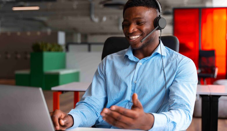 "Smiling young African man wearing a headset, sitting at a desk in a modern office, engaged in an online meeting on his laptop.