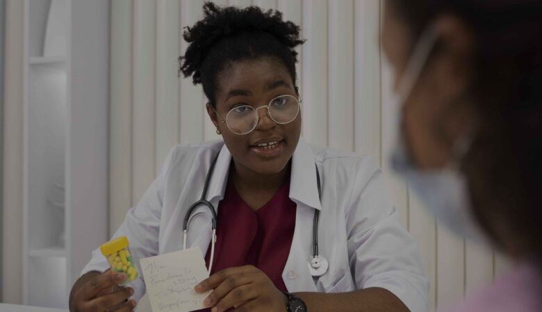 A female doctor wearing glasses and a stethoscope is seated at a desk, holding a small pill bottle in one hand and a prescription note in the other. She is explaining something to a patient sitting across from her, who is partially visible and wearing a face mask. An open notebook lies on the desk in front of the doctor, creating a professional and caring atmosphere in a medical setting.